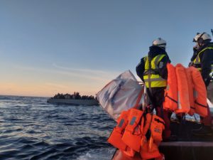 Im Hintergrund ist ein überfülltes Boot auf dem offenen Meer sichtbar. Im Vordergrund ist ein Ausschnitt von einem kleinen Rettungsboot mit zwei Personen mit Schutzausrüstung und mehreren organgenen Schwimmwesten an Board, abgebildet.