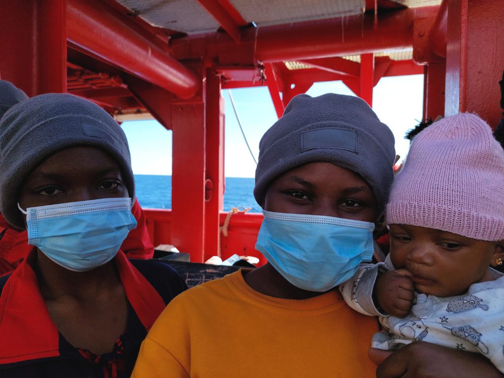 Portrait von zwei Frauen mit einem Baby auf dem Arm. Sie tragen alle drei Mützen und die zwei Frauen blaue medizinische Masken über Mund und Nase. Im Hintergrund ist die rote Gerüststruktur vom Schiff zu sehen und das Meer. 