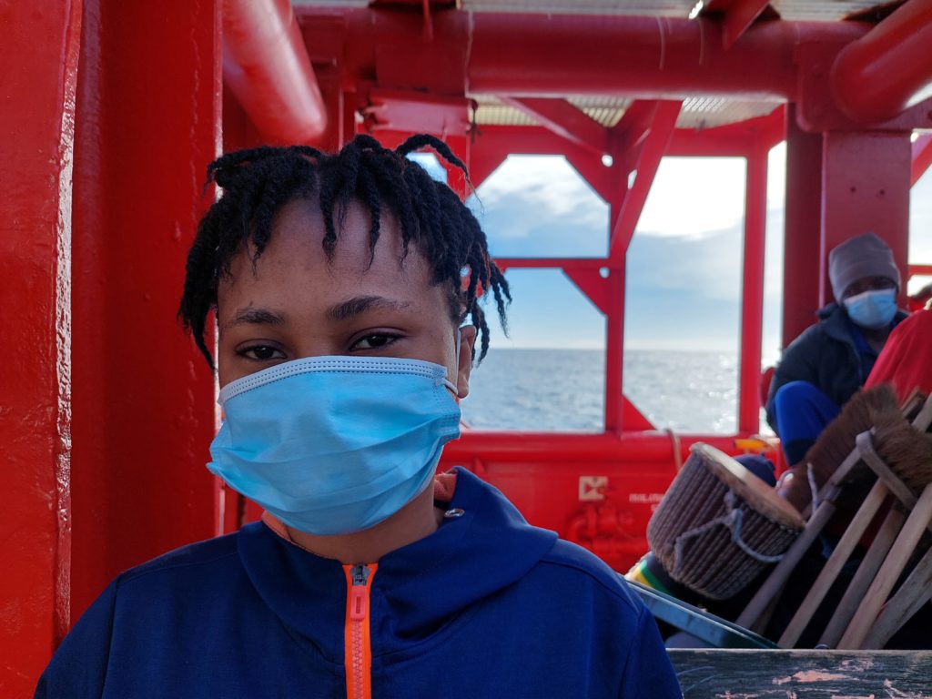 Portrait einer Frau, die frontal in die Kamera guckt. Sie trägt eine blaue medizinische Maske über Mund und Nase. Im Hintergrund ist die Strutur des Schiffes zu erkennen, mit einem Ausblick zum Meer.