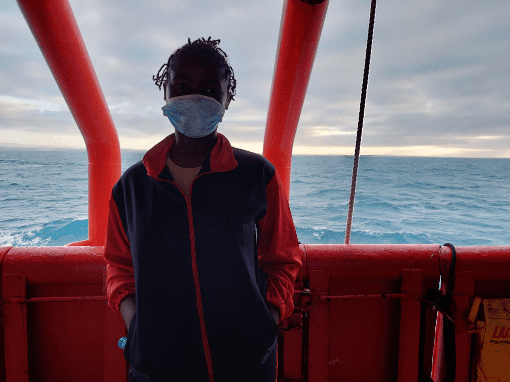 Foto von einer Frau vor dem roten Schiffsgerüst und dem Meer. Die Frau ist bis zur Hüfte aufwerts zu sehen. Sie trägt eine blaue medizinische Maske und eine blau-rote Jacke. Sie hat die Hände in den Jackentaschen.